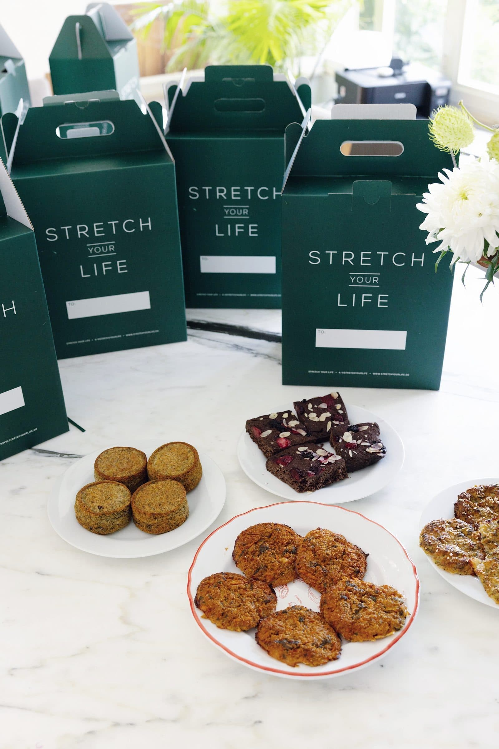 Mermer tezgâhta yeşil Stretch Your Life kutuları; önlerinde tabaklarda muffin, brownie ve diğer ürünler